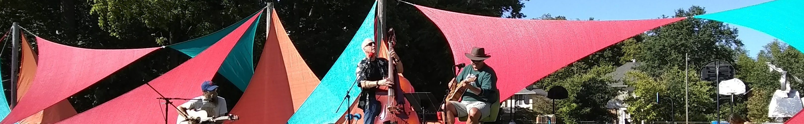 Two guitarists and an upright bass player perform in front of the colorful sails sculpture on a beautiful day.